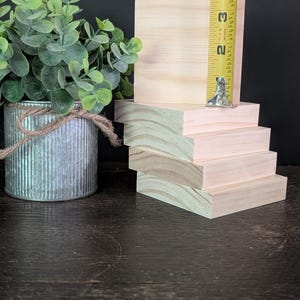 May include: A stack of unfinished, light-colored wooden blocks with a measuring tape showing a height of approximately 4 inches. A small metal clip holds the tape in place. A decorative plant in a galvanized metal pot sits to the left.