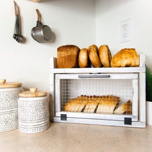 May include: White wooden bread box with a wire mesh front. The box has two shelves, the top shelf holds a loaf of bread and several rolls, and the bottom shelf holds sliced bread. Two ceramic canisters with wooden lids are in the foreground.