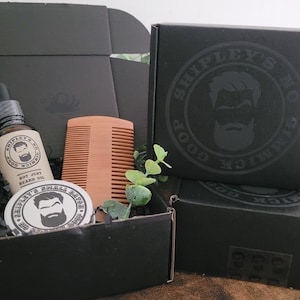 May include: A black gift box with a wooden comb, a small jar of beard balm, and a bottle of beard oil. The box is decorated with a black and white logo that says "Shipley's Good Ol' Fashioned Goods".