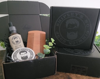 May include: A black gift box with a wooden comb, a small jar of beard balm, and a bottle of beard oil. The box is decorated with a black and white logo that says "Shipley's Good Ol' Fashioned Goods".