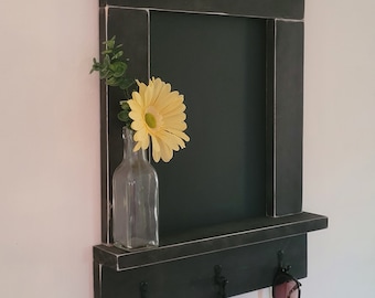 May include: Black wooden key holder with a chalkboard, a shelf, and three hooks. A small glass vase with a yellow flower sits on the shelf.