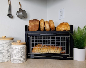 May include: A black bread box with a wire mesh door and a shelf for storing bread. The bread box is on a countertop with two ceramic canisters and a potted plant.