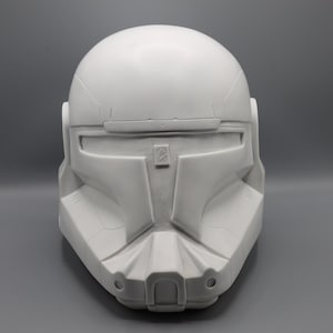 May include: A white, unpainted helmet with a visor and detailed facial features. The helmet has a rounded top and a rectangular visor opening. The design includes angular cheek pieces and a central, vertical panel. The overall aesthetic suggests a futuristic or science fiction theme.