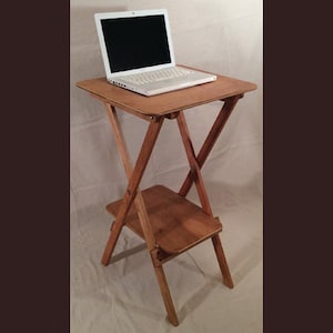 May include: A wooden laptop stand with a light-colored laptop on top. The stand is made of light brown wood and has a lower shelf. The stand is approximately 30 inches tall.