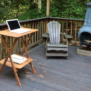 May include: A wooden folding table with a laptop on the top shelf and a wire basket on the bottom shelf. The table is on a wooden deck with a chair and a fire pit in the background.