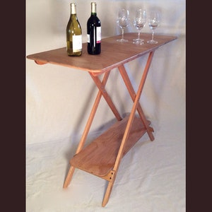May include: A wooden folding table with a shelf, two bottles of wine, and four wine glasses. The table is made of light wood and has a natural finish.