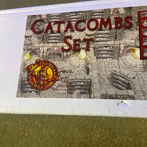 May include: Close-up of a box with the words "CATACOMS SET" in red lettering. The box features a stone-like background and a logo with a dwarf figure. The text "DWARVEN FORGE" is also visible.