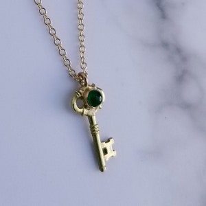 Op de afbeelding: Een gouden sleutelketting met een groene edelsteen. De sleutel heeft een gedetailleerd ontwerp en de edelsteen is in de bovenkant van de sleutel gezet.