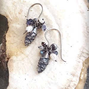 May include: A pair of silver earrings with a pine cone charm and a cluster of white and purple beads.