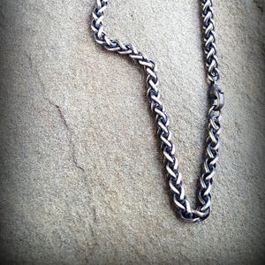 Puede incluir: Un collar de cadena de plata con un dise&ntilde;o trenzado. La cadena est&aacute; sobre una superficie gris texturizada.
