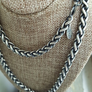 Puede incluir: Un collar de cadena de plata con un dise&ntilde;o trenzado. La cadena se muestra en un maniqu&iacute; de tela marr&oacute;n.