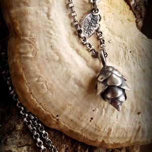 May include: A silver chain necklace with a small silver pine cone pendant. The necklace is on a light brown surface.