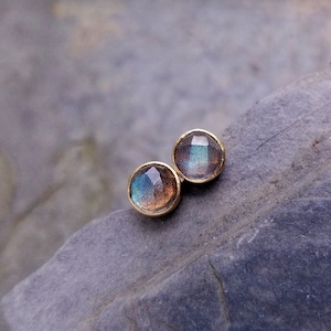 May include: A pair of round stud earrings with gold settings. Each earring features a faceted, iridescent gemstone with hues of blue, green, and brown. The earrings are displayed on a textured, gray stone surface.