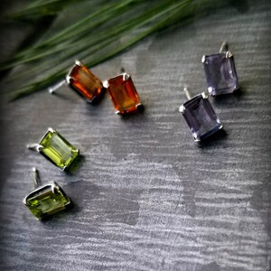 Peut inclure: Quatre paires de boucles d'oreilles avec des pierres précieuses de différentes couleurs. Les boucles d'oreilles sont en argent et ont une forme rectangulaire. Les pierres précieuses sont vertes, orange et violettes.