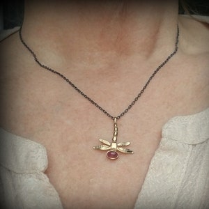 Puede incluir: Un collar con colgante de libélula dorado con una cadena oscura. La libélula tiene alas detalladas y una gema roja en el centro. El collar se lleva sobre una camisa blanca.
