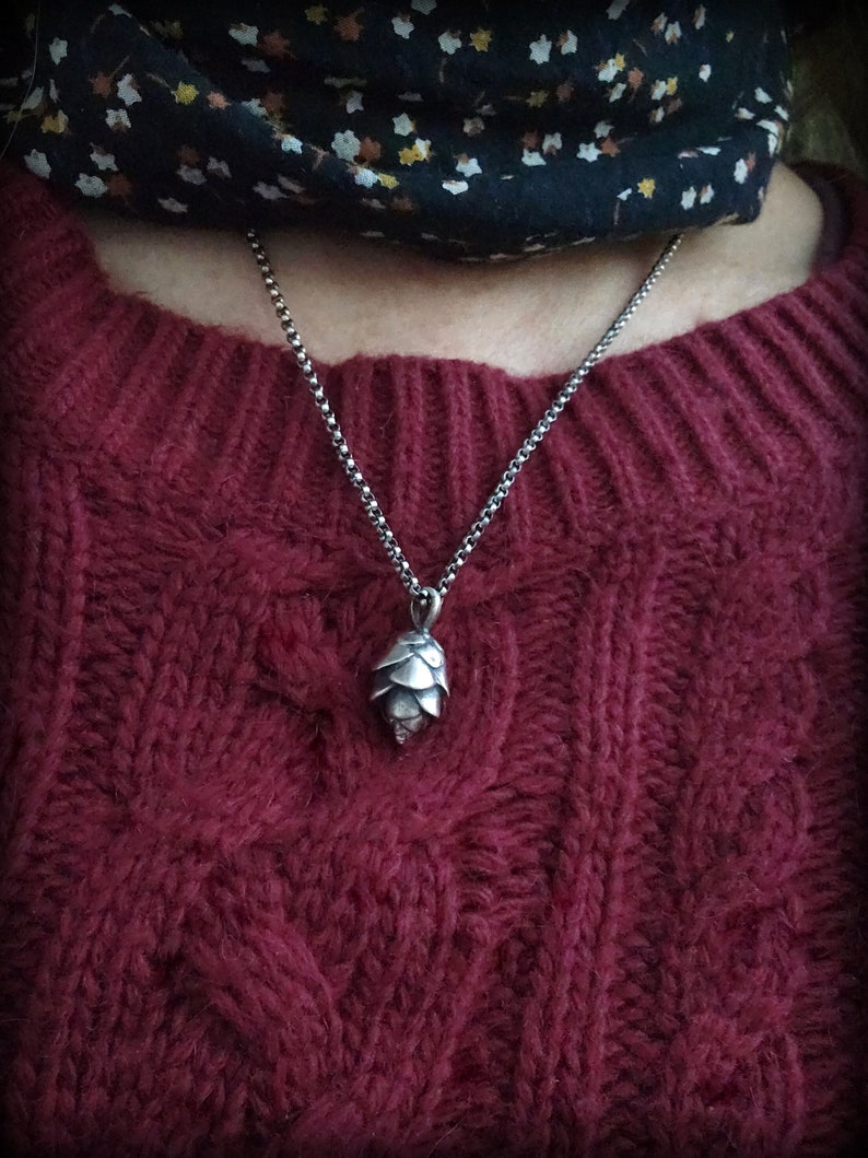 May include: A silver chain necklace with a silver pine cone pendant. The necklace is worn over a burgundy sweater.