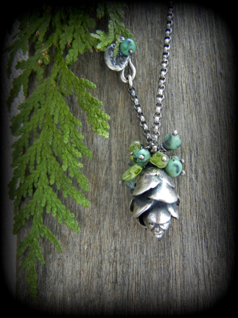 May include: A silver necklace with a pine cone pendant and green gemstone accents. The necklace is on a wooden background with a green leafy branch.