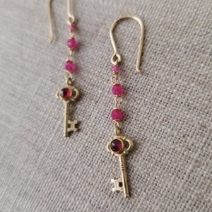 Könnte beinhalten: Ein Paar goldfarbene Ohrringe mit einem Schlüsseldesign. Jeder Ohrring hat einen Schlüsselanhänger mit einem roten Edelstein und ist mit kleinen, rosa Perlen verziert. Die Ohrringe hängen an einem gebogenen Haken.