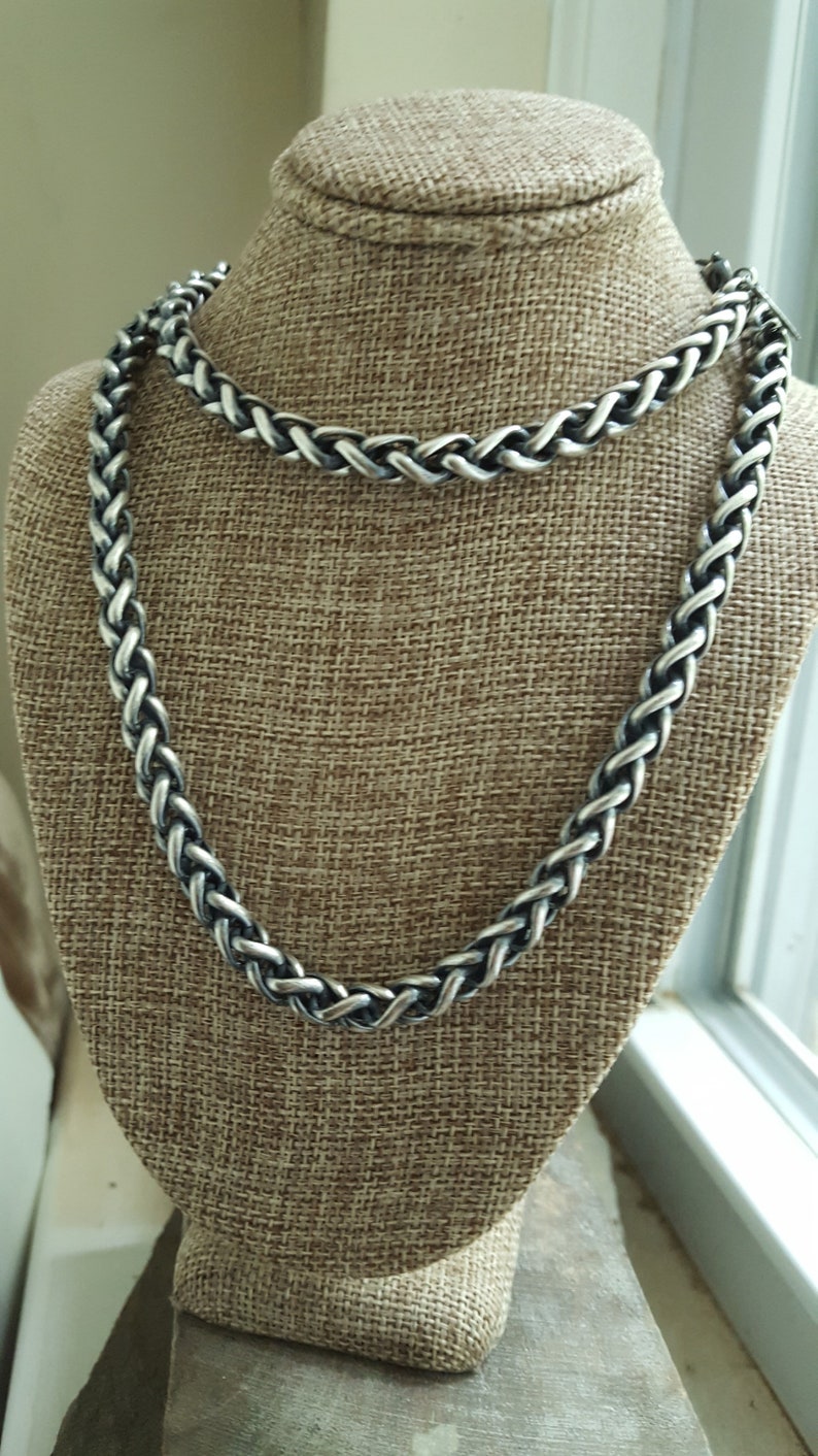 Puede incluir: Un collar de cadena de plata con un dise&ntilde;o trenzado. El collar se muestra en un maniqu&iacute; de tela marr&oacute;n.