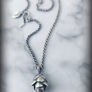 May include: A silver chain necklace with a silver pine cone pendant. The necklace is on a white marble background.