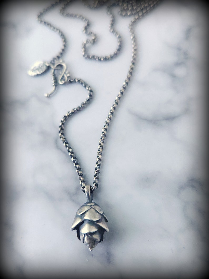 May include: A silver chain necklace with a silver pine cone pendant. The necklace is on a white marble background.