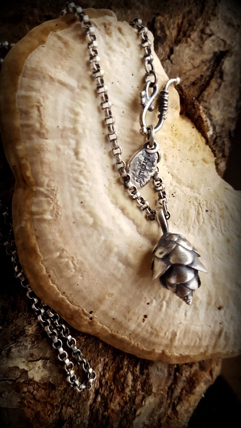 May include: A silver chain necklace with a small silver pine cone pendant. The necklace is on a light brown surface.