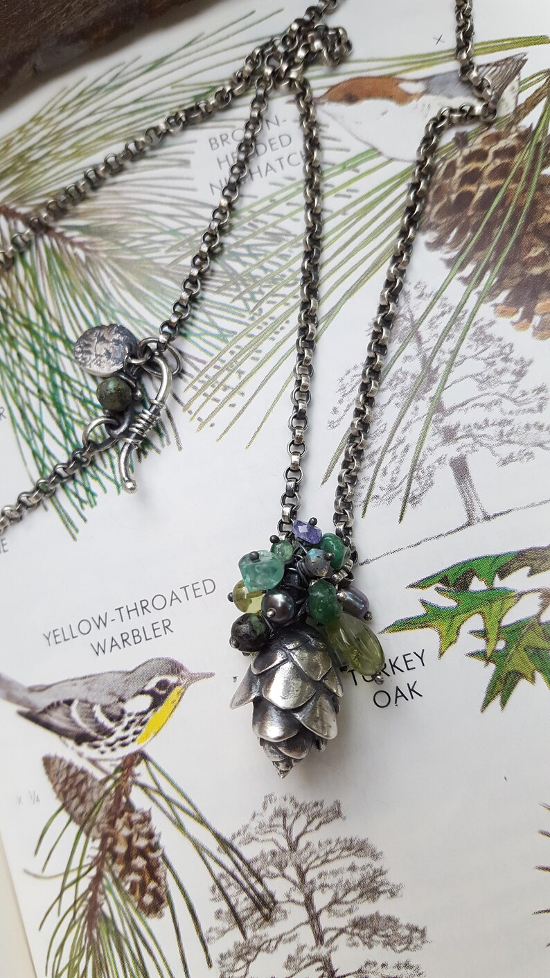 May include: A silver chain necklace with a silver pine cone pendant adorned with green and purple gemstones. The necklace is displayed on a book page with illustrations of a bird and a tree.