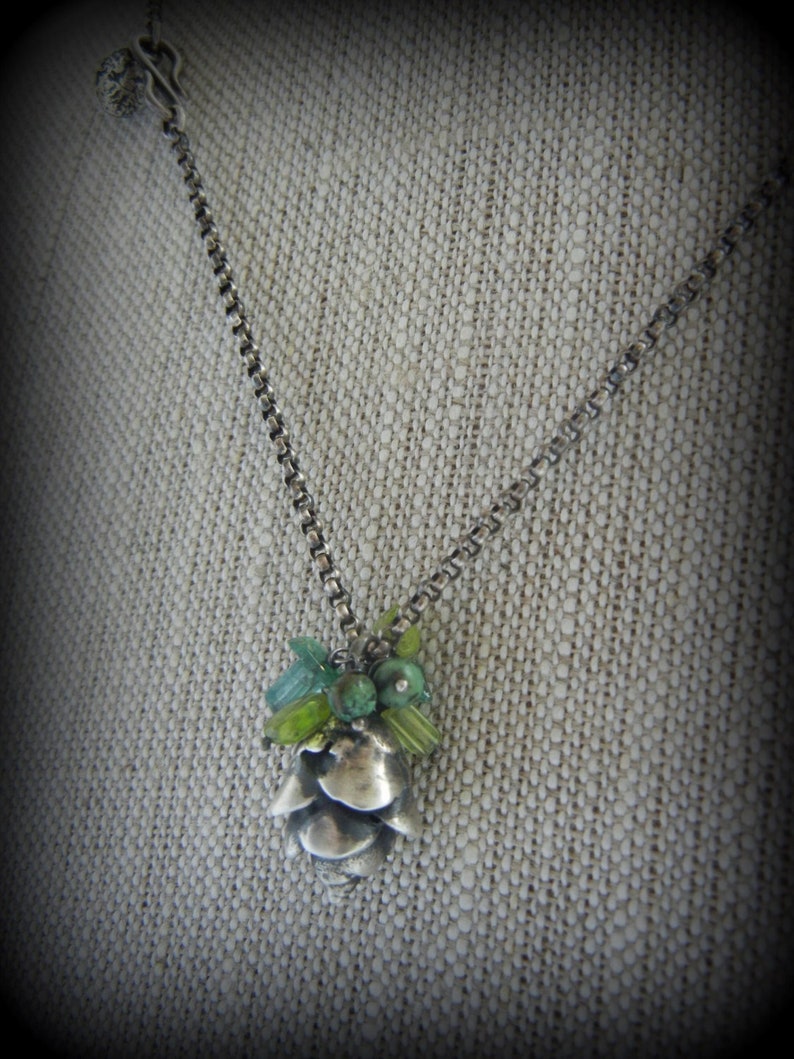 May include: A silver chain necklace with a silver pine cone pendant adorned with green and blue gemstones.