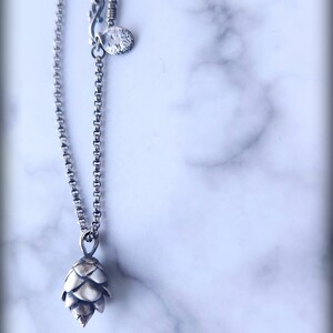 May include: A silver chain necklace with a small silver pine cone pendant. The chain has a lobster clasp and a small silver disc charm.