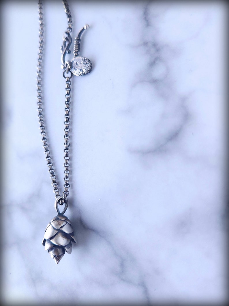 May include: A silver chain necklace with a small silver pine cone pendant. The chain has a lobster clasp and a small silver disc charm.