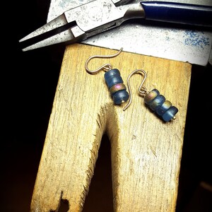 May include: A pair of earrings with blue and gold beads hanging from gold hooks. The earrings are on a wooden surface with a pair of pliers in the background.