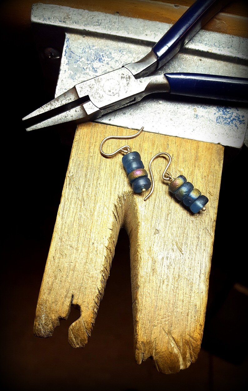 May include: A pair of earrings with blue and gold beads hanging from gold hooks. The earrings are on a wooden surface with a pair of pliers in the background.