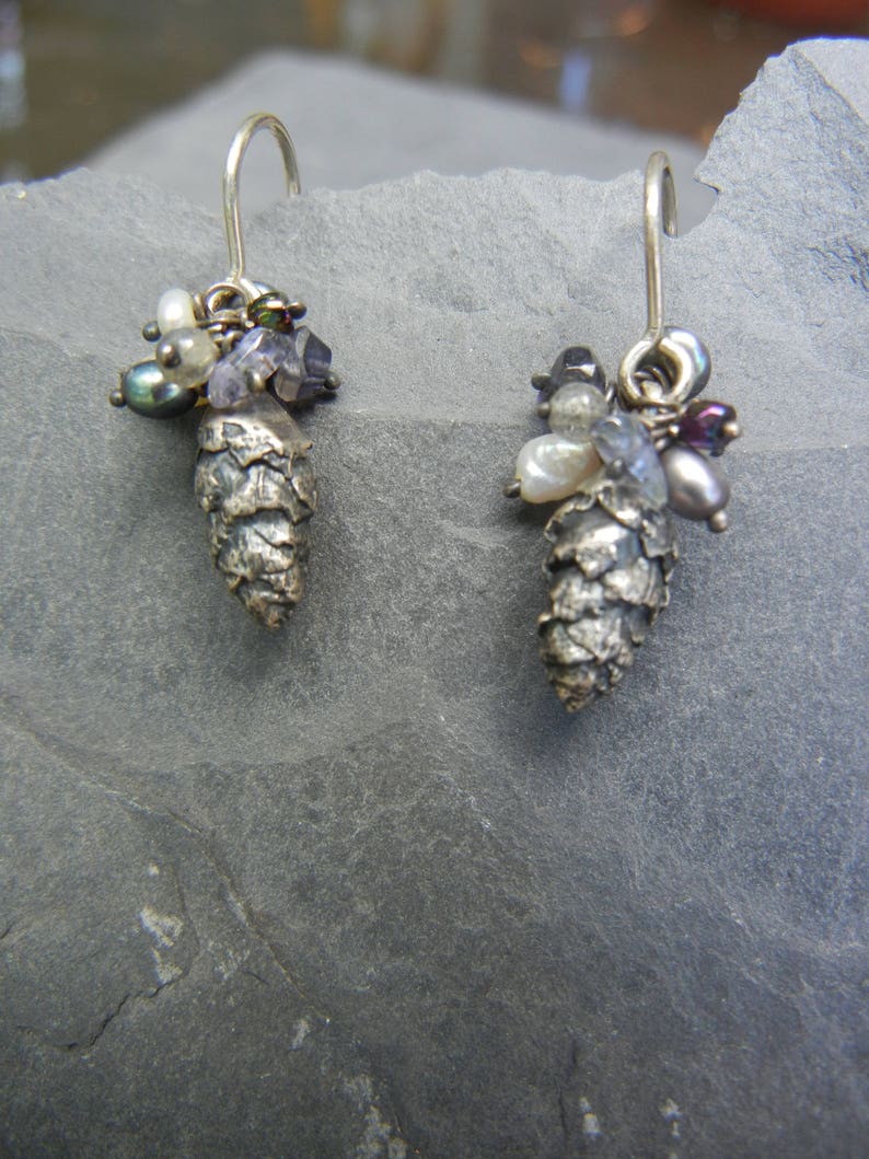 May include: A pair of silver earrings with a pine cone design. Each earring is adorned with a cluster of small, colorful beads, including white, purple, and black.