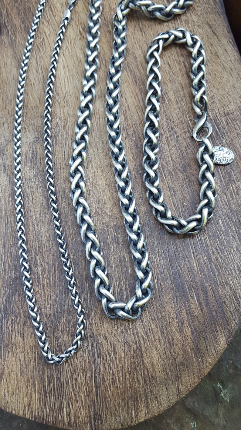 Puede incluir: Cuatro cadenas de plata de diferentes anchos y longitudes, mostradas sobre una superficie de madera. Las cadenas est&aacute;n hechas con un dise&ntilde;o trenzado.