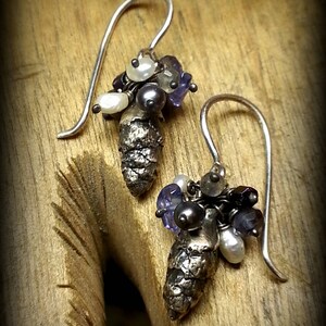 May include: A pair of silver earrings with a pine cone charm and a cluster of purple and white beads.