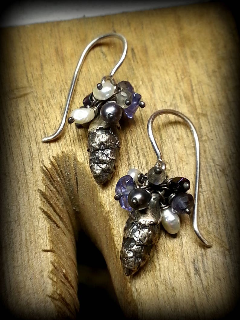 May include: A pair of silver earrings with a pine cone charm and a cluster of purple and white beads.