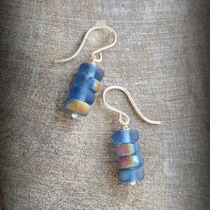 May include: A pair of silver dangle earrings with blue and iridescent glass beads. The beads are stacked on top of each other and have a slightly rounded shape.
