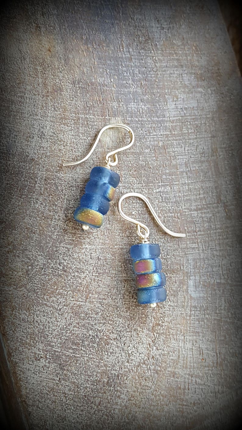 May include: A pair of silver dangle earrings with blue and iridescent glass beads. The beads are stacked on top of each other and have a slightly rounded shape.