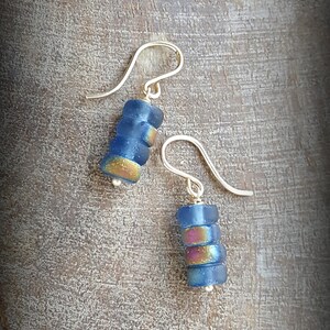 May include: A pair of silver dangle earrings with blue and iridescent glass beads. The beads are stacked on top of each other and have a slightly rounded shape.