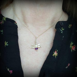 Peut inclure: Un collier en or avec un pendentif en forme de libellule orné d'une pierre précieuse violette. La libellule a des ailes détaillées et un corps texturé.