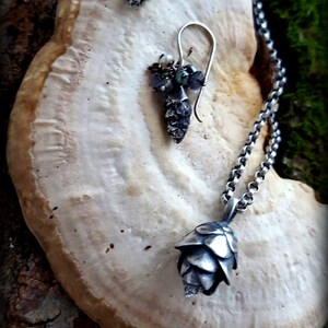 May include: A silver necklace with a pine cone pendant and matching earrings. The earrings feature a cluster of small, iridescent gemstones. The jewelry is displayed on a light brown, textured surface.