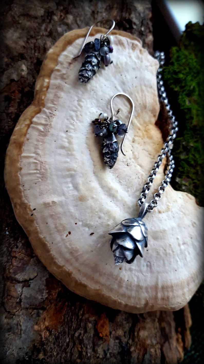 May include: A silver necklace with a pine cone pendant and matching earrings. The earrings feature a cluster of small, iridescent gemstones. The jewelry is displayed on a light brown, textured surface.