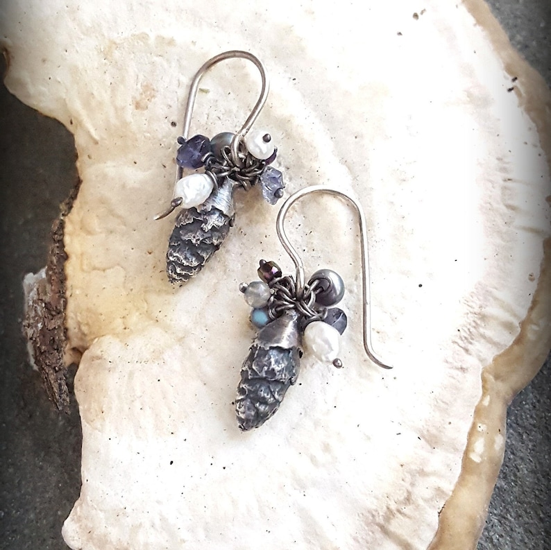 May include: A pair of silver earrings with a pine cone charm and a cluster of white and purple beads.