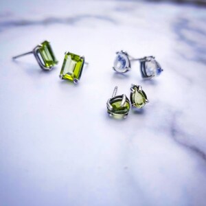 May include: Three pairs of earrings with green and white gemstones. The earrings are silver and have a variety of shapes, including square, oval, and teardrop.