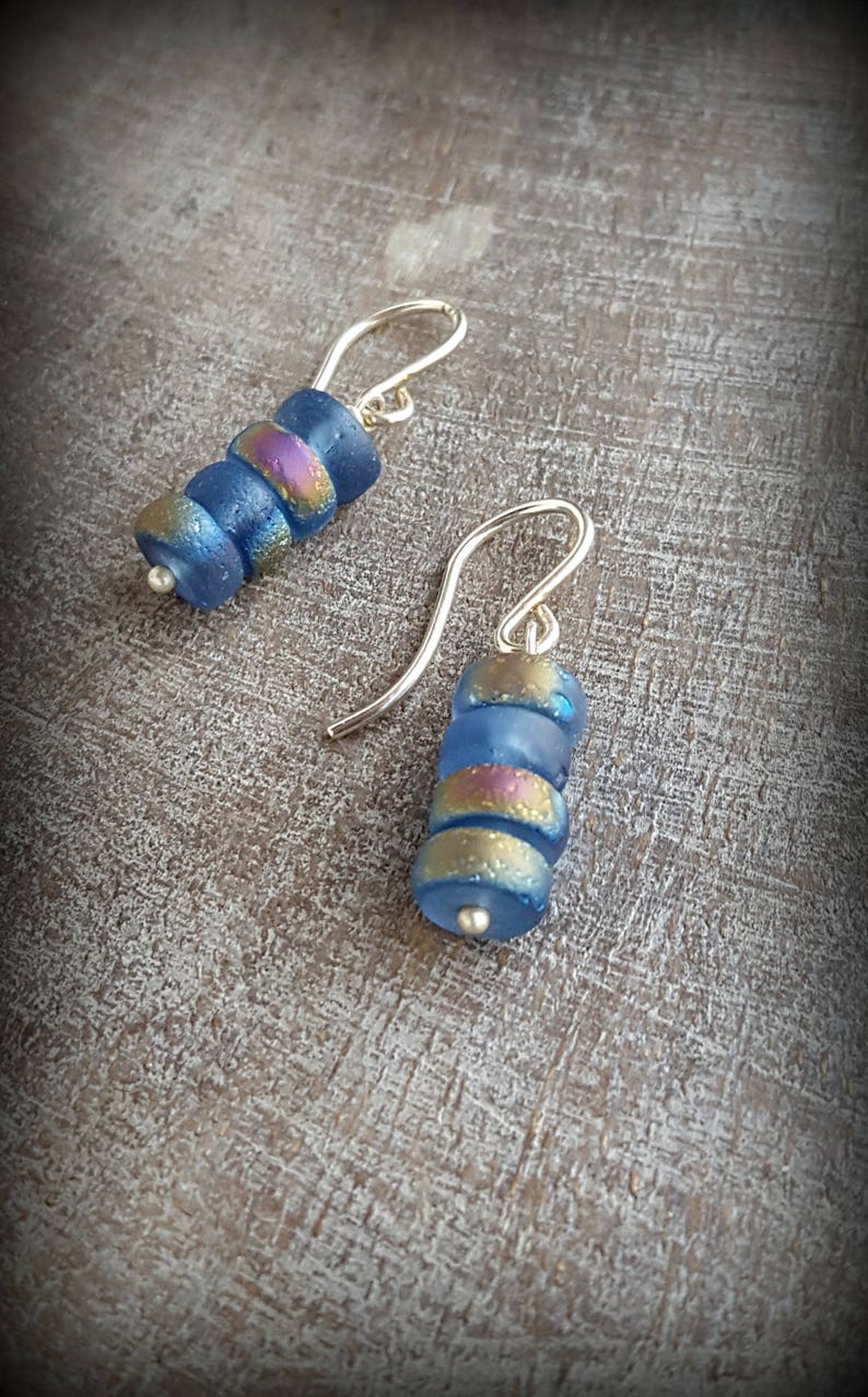 May include: A pair of silver dangle earrings with blue and iridescent glass beads. The beads are stacked on top of each other and have a slightly textured surface.