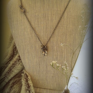 May include: A silver chain necklace with a small, intricate pendant made of metal and beads. The necklace is displayed on a beige fabric mannequin with a small bouquet of white flowers in a glass bottle.
