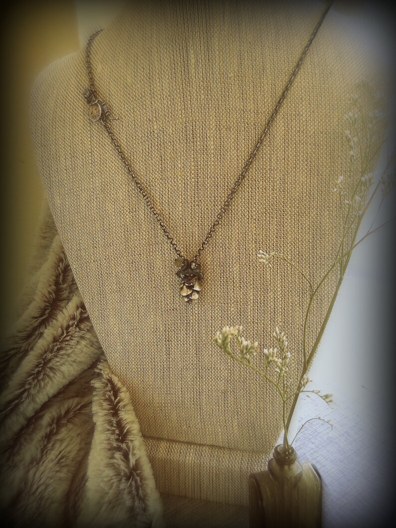 May include: A silver chain necklace with a small, intricate pendant made of metal and beads. The necklace is displayed on a beige fabric mannequin with a small bouquet of white flowers in a glass bottle.