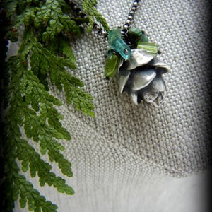 May include: A silver pinecone pendant necklace with green and blue glass beads. The necklace is displayed on a light brown fabric background.