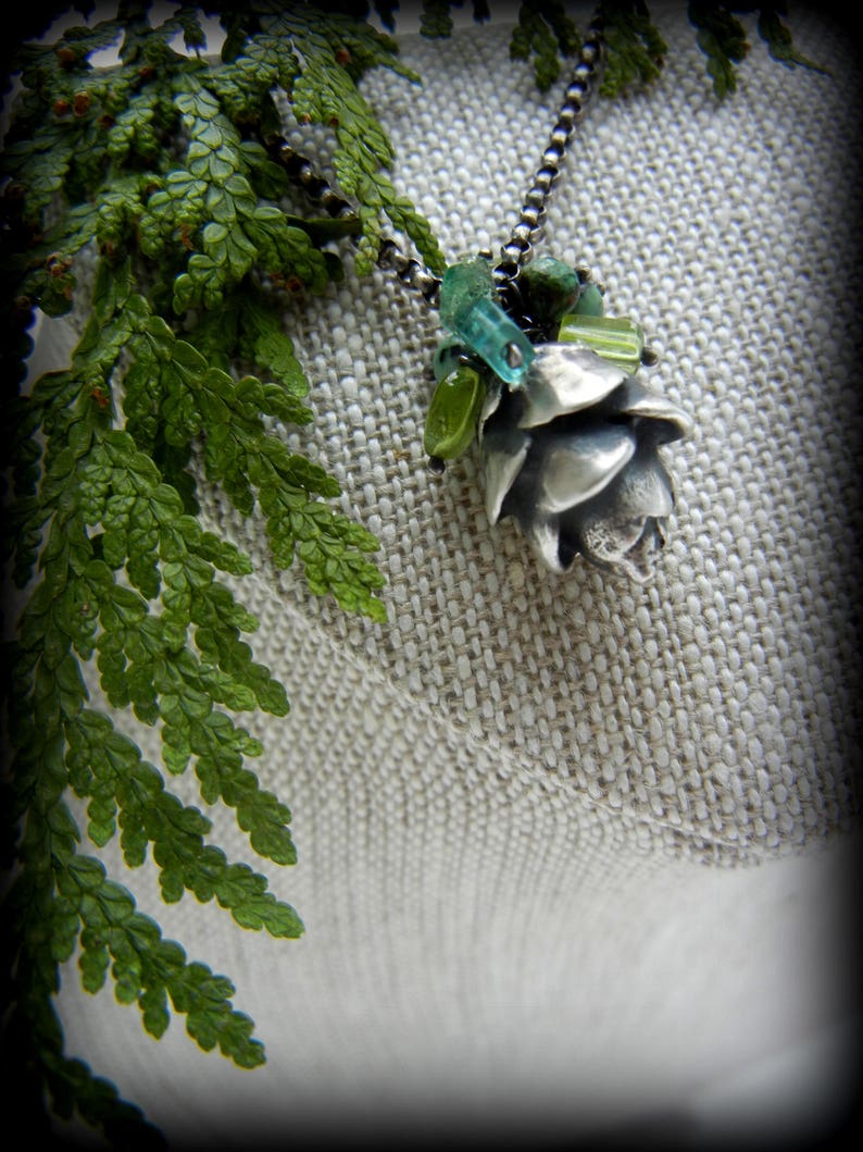 May include: A silver pinecone pendant necklace with green and blue glass beads. The necklace is displayed on a light brown fabric background.