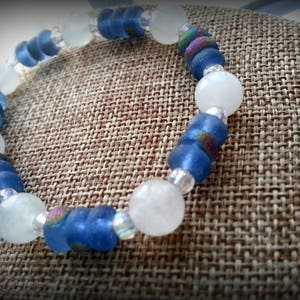 May include: A blue and white beaded bracelet with iridescent accents. The bracelet is made of glass beads and is displayed on a brown fabric background.